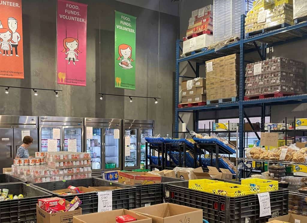 An inside look at the Central Okanagan Community Food Bank. There are four commercial freezers, palettes stacked in commercial-grade storage shelves, and a collection of big plastic pins featuring different non-perishables.